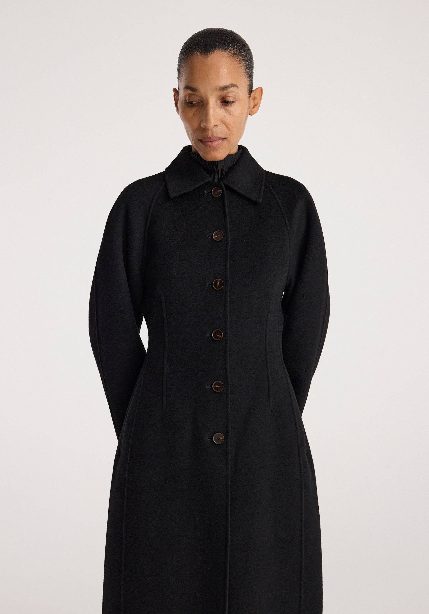 Woman wearing a sleek black Double-faced elongated sculptural coat | black with button closure and sculpted long sleeves against a plain light background.