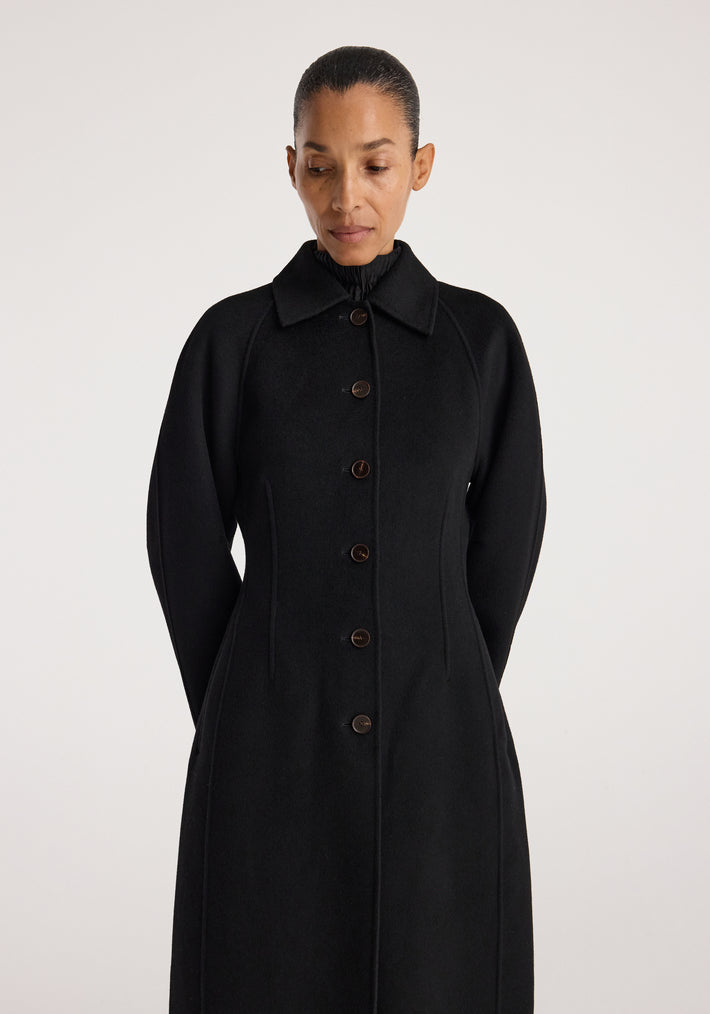 Woman wearing a sleek black Double-faced elongated sculptural coat | black with button closure and sculpted long sleeves against a plain light background.
