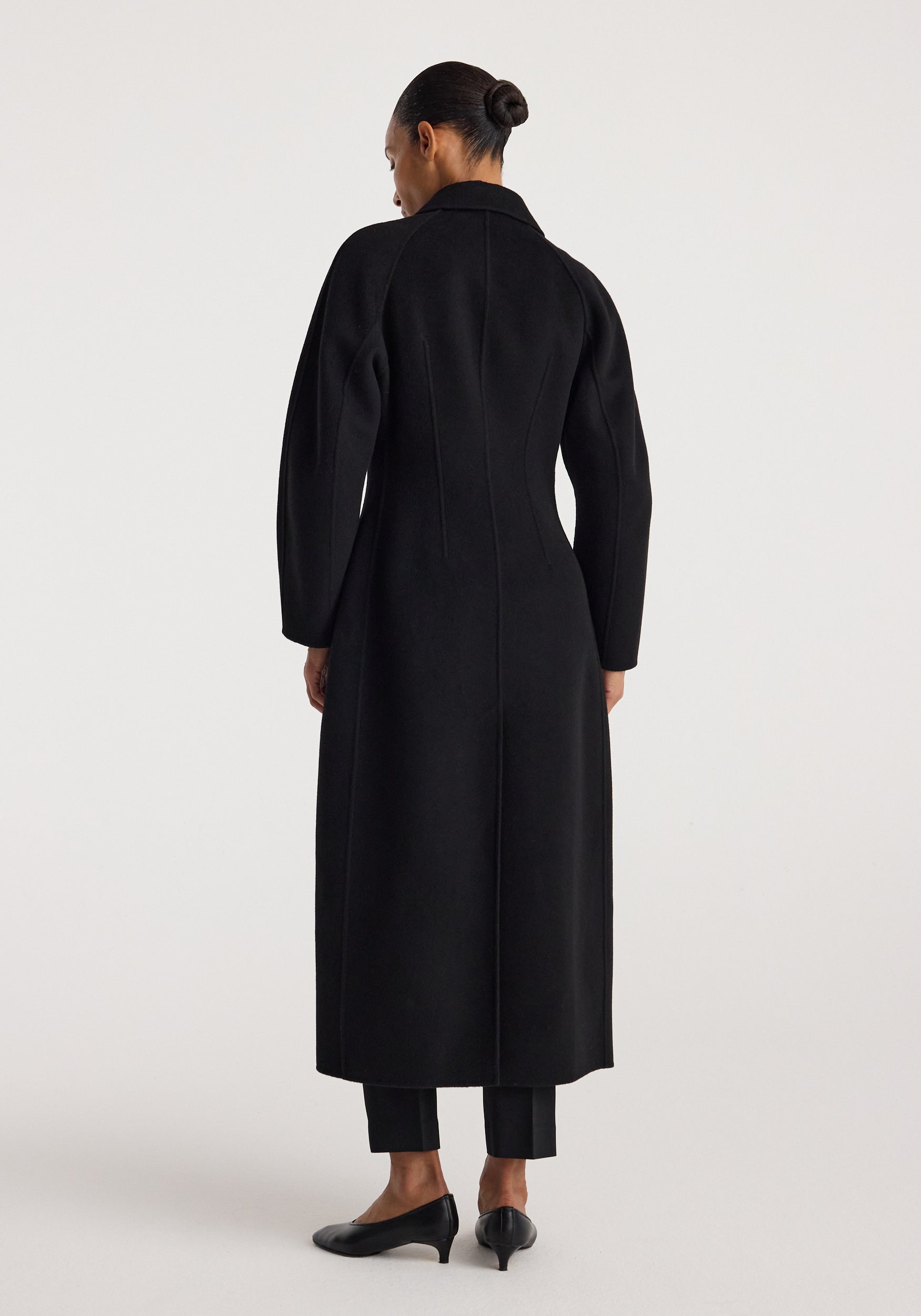 Woman wearing a sleek double-faced elongated sculptural coat | black, standing with her back to the camera, showcasing the coat’s tailored fit and long silhouette.