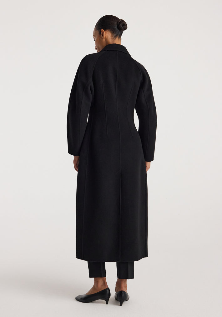 Woman wearing a sleek double-faced elongated sculptural coat | black, standing with her back to the camera, showcasing the coat’s tailored fit and long silhouette.