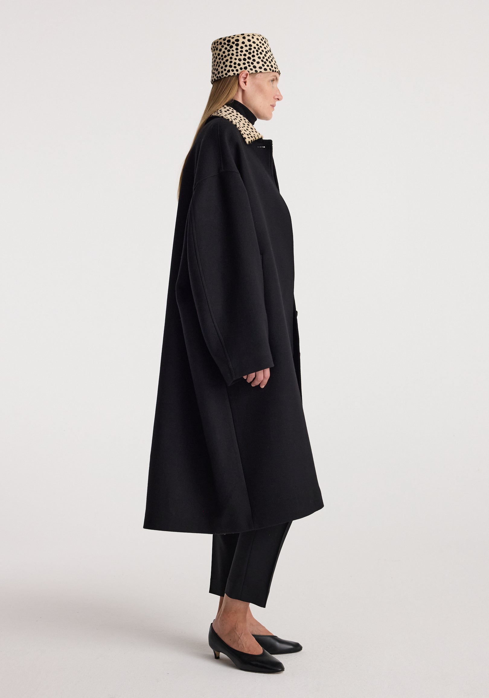 Side profile of a woman wearing a black cheetah collar wool coat | black paired with matching cheetah print headwear and black shoes against a plain background.