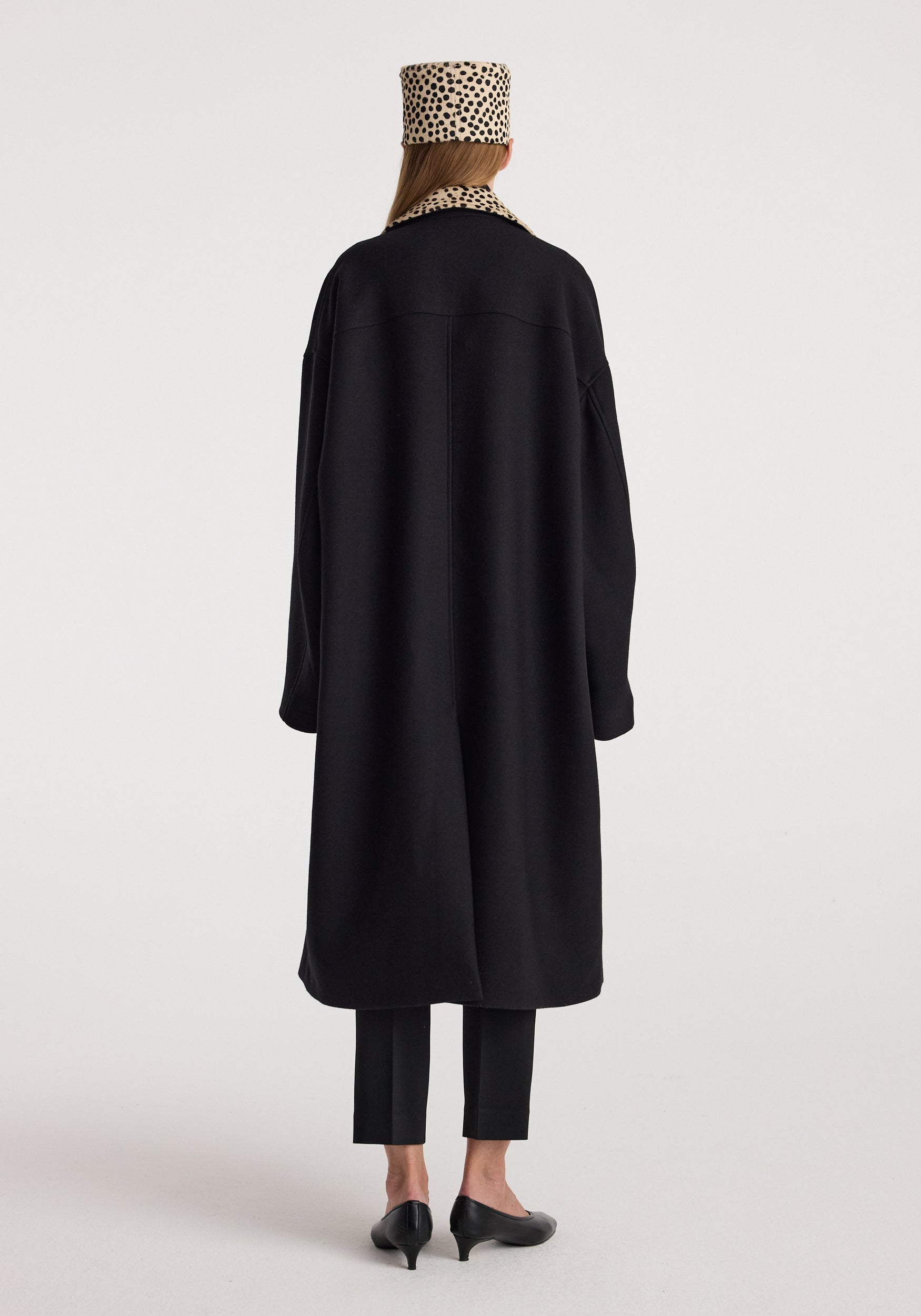 Woman wearing a long black coat with a cheetah-print collar, paired with black slim trousers, black low-heeled shoes, and a matching cheetah-print hat, shown from the back, featuring the Cheetah collar wool coat | black.
