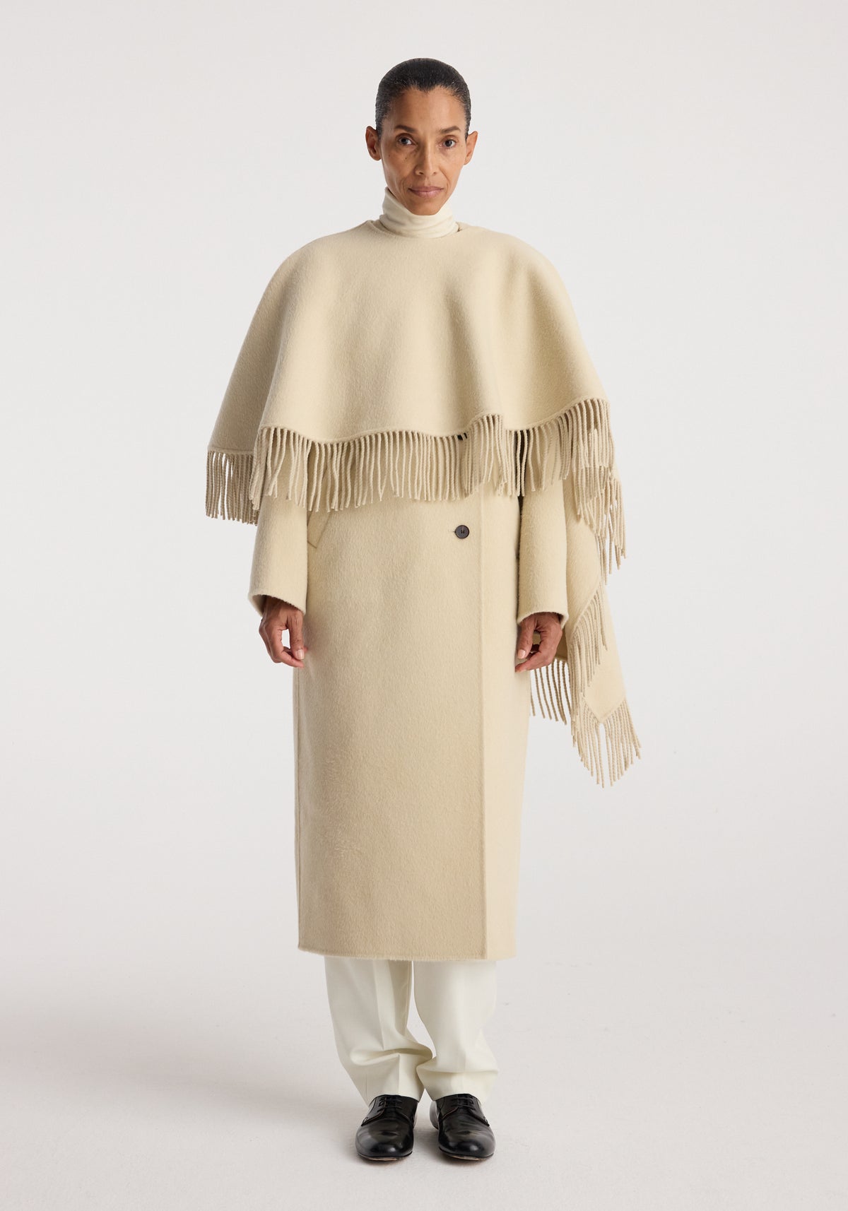 Woman wearing a cream-colored double-faced fringe coat | butter with long sleeves and fringe detailing, paired with white pants and black shoes against a plain background.