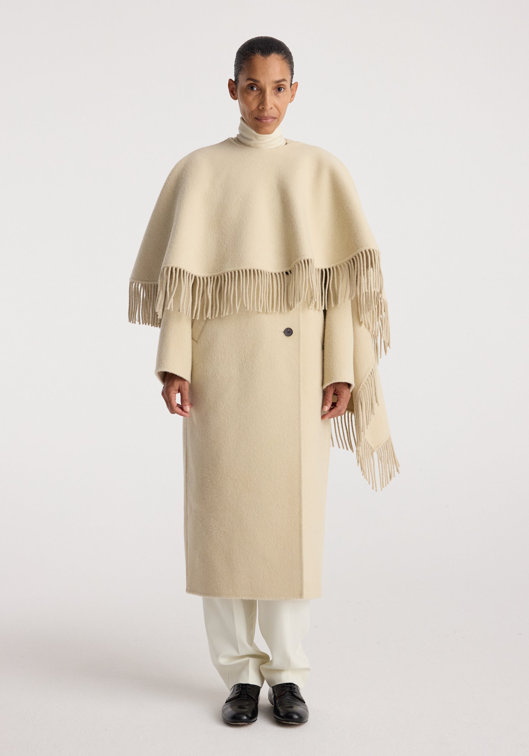 Woman wearing a cream-colored double-faced fringe coat | butter with long sleeves and fringe detailing, paired with white pants and black shoes against a plain background.
