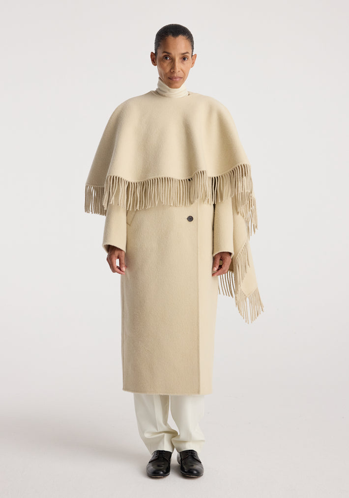 Woman wearing a cream-colored double-faced fringe coat | butter with long sleeves and fringe detailing, paired with white pants and black shoes against a plain background.