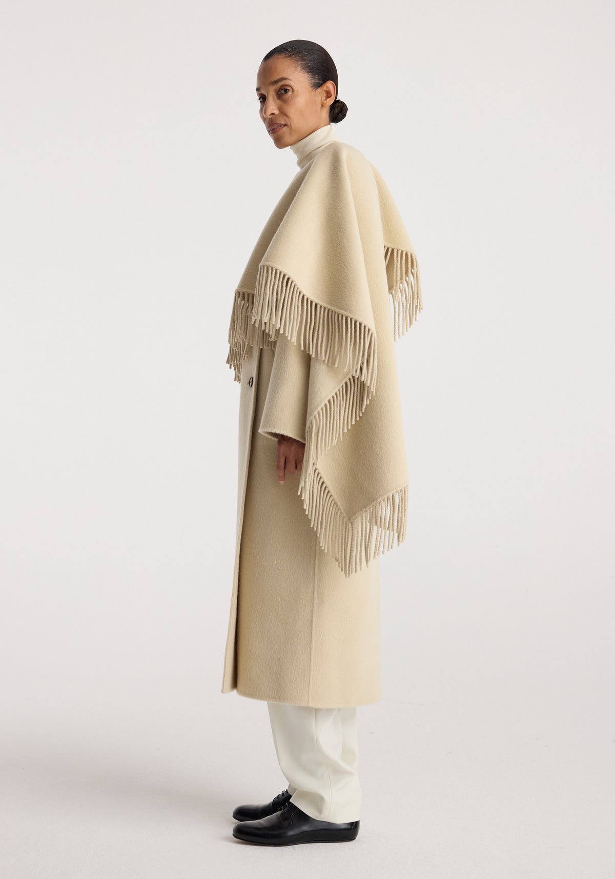 Woman wearing a light beige double-faced fringe coat | butter with long fringed edges, styled over white pants and black shoes against a plain background.