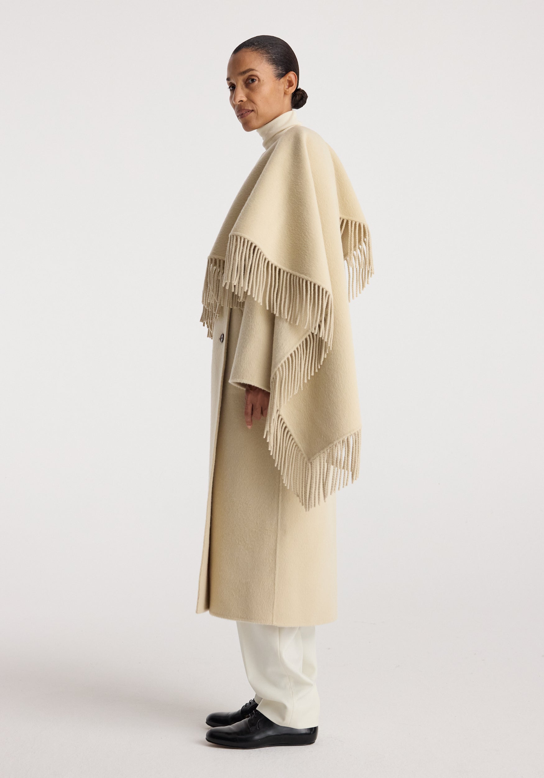 Woman wearing a light beige double-faced fringe coat | butter with long fringed edges, styled over white pants and black shoes against a plain background.