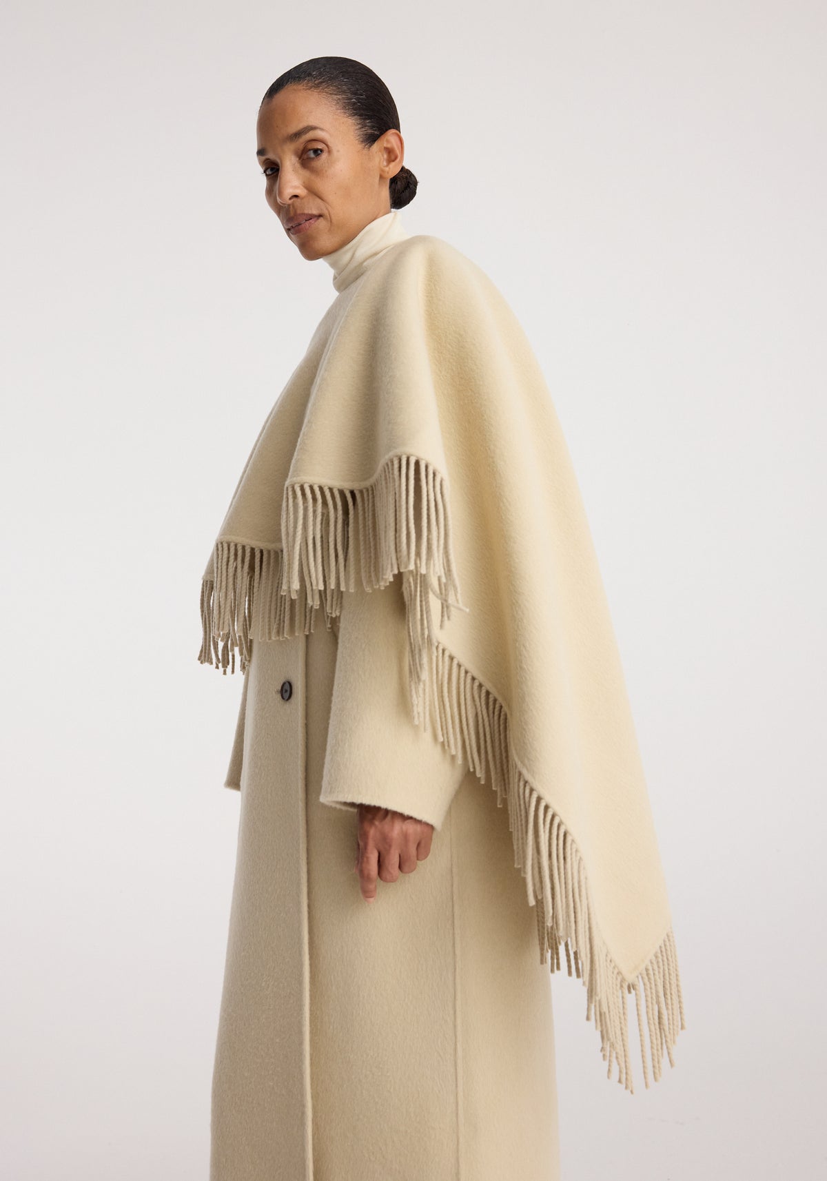 Woman wearing a light cream-colored double-faced fringe coat | butter with a high collar and draped fringe detailing along the edges, standing against a plain background.