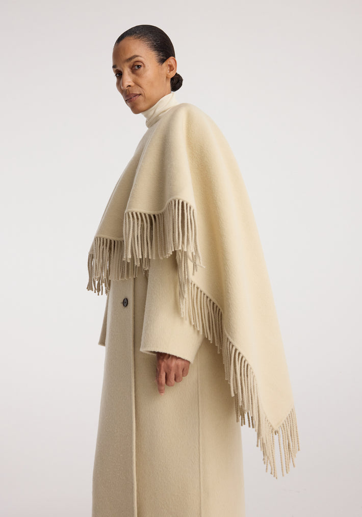 Woman wearing a light cream-colored double-faced fringe coat | butter with a high collar and draped fringe detailing along the edges, standing against a plain background.