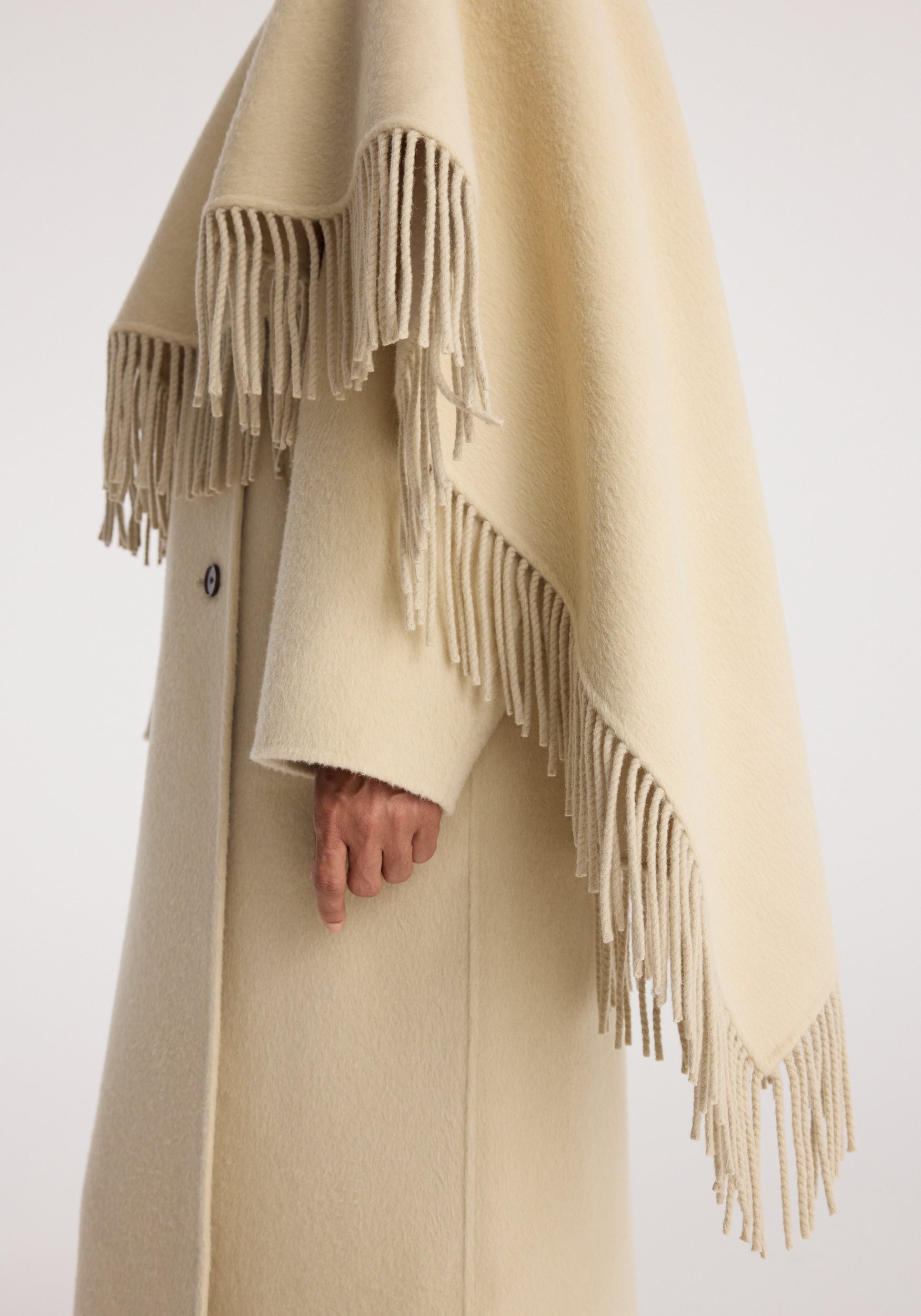 Close-up of a person wearing a soft cream-colored Double-faced fringe coat | butter with long fringe details along the edges and a buttoned sleeve visible.