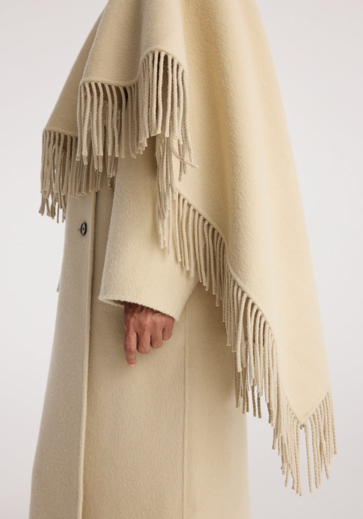 Close-up of a person wearing a soft cream-colored Double-faced fringe coat | butter with long fringe details along the edges and a buttoned sleeve visible.
