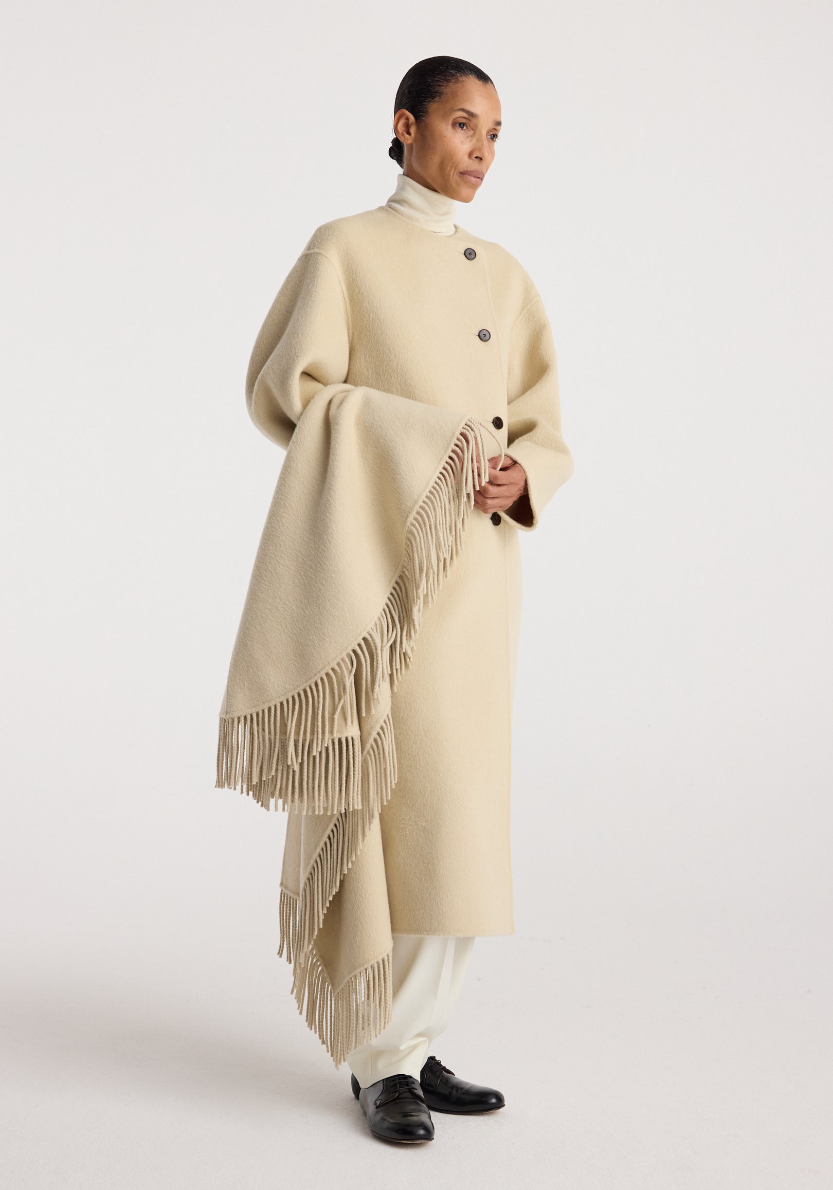 A person wearing a cream-colored Double-faced fringe coat | butter with a high collar, featuring large buttons down the front and long fringes along one side, paired with white pants and black shoes.