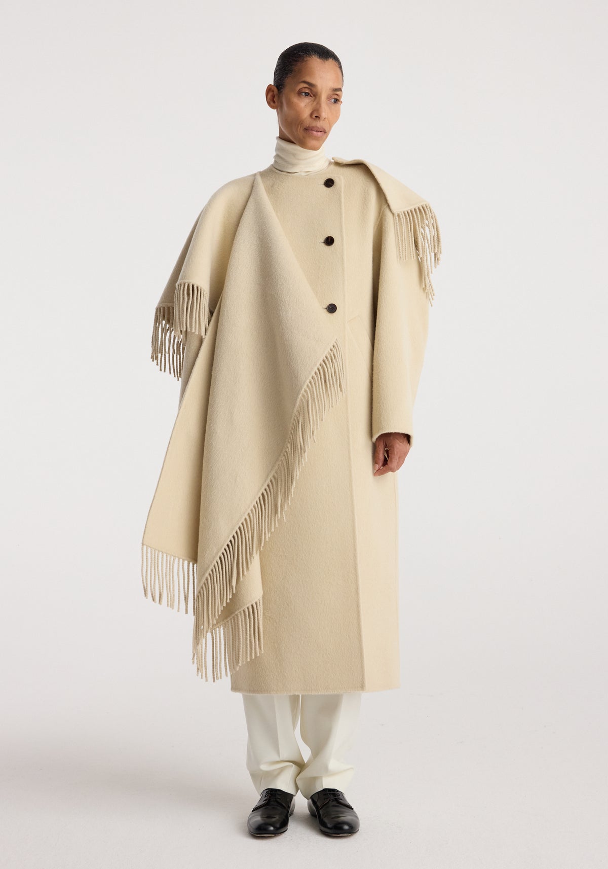 Woman wearing a cream-colored Double-faced fringe coat | butter with large buttons, fringed edges, and a draped design over a turtleneck and white pants.