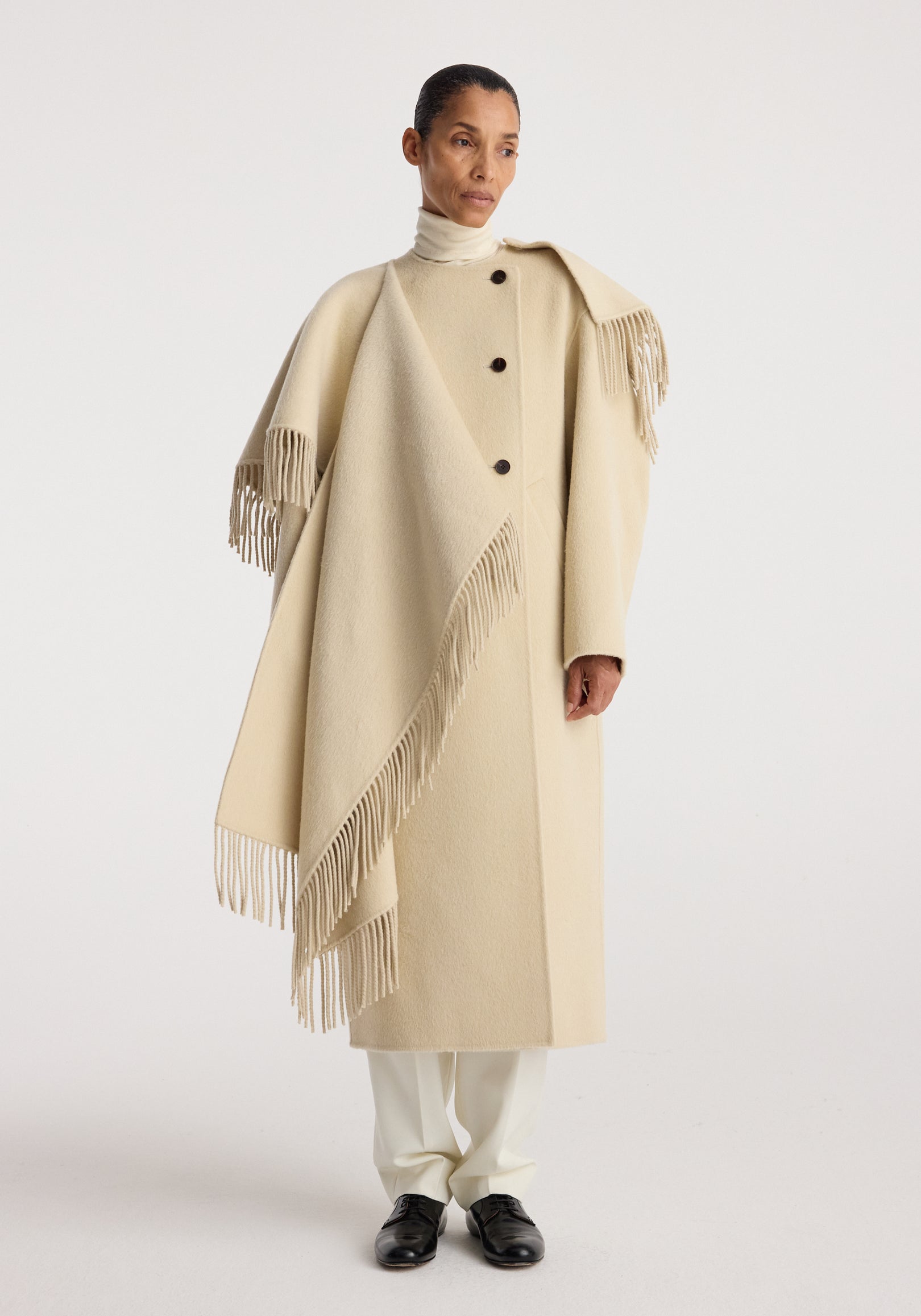 Woman wearing a cream-colored Double-faced fringe coat | butter with large buttons, fringed edges, and a draped design over a turtleneck and white pants.