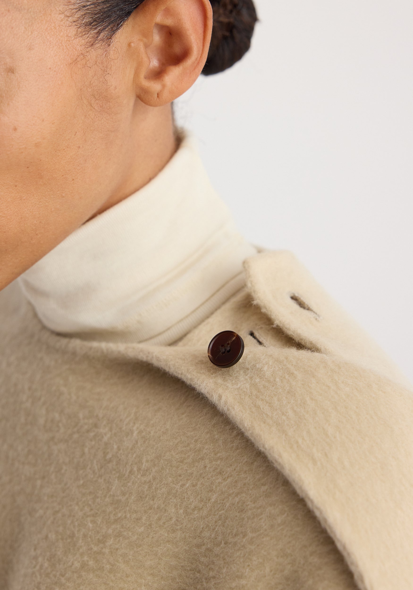 Close-up of a person wearing a cream-colored Double-faced fringe coat | butter with a dark brown button detail on the shoulder over a white turtleneck sweater.