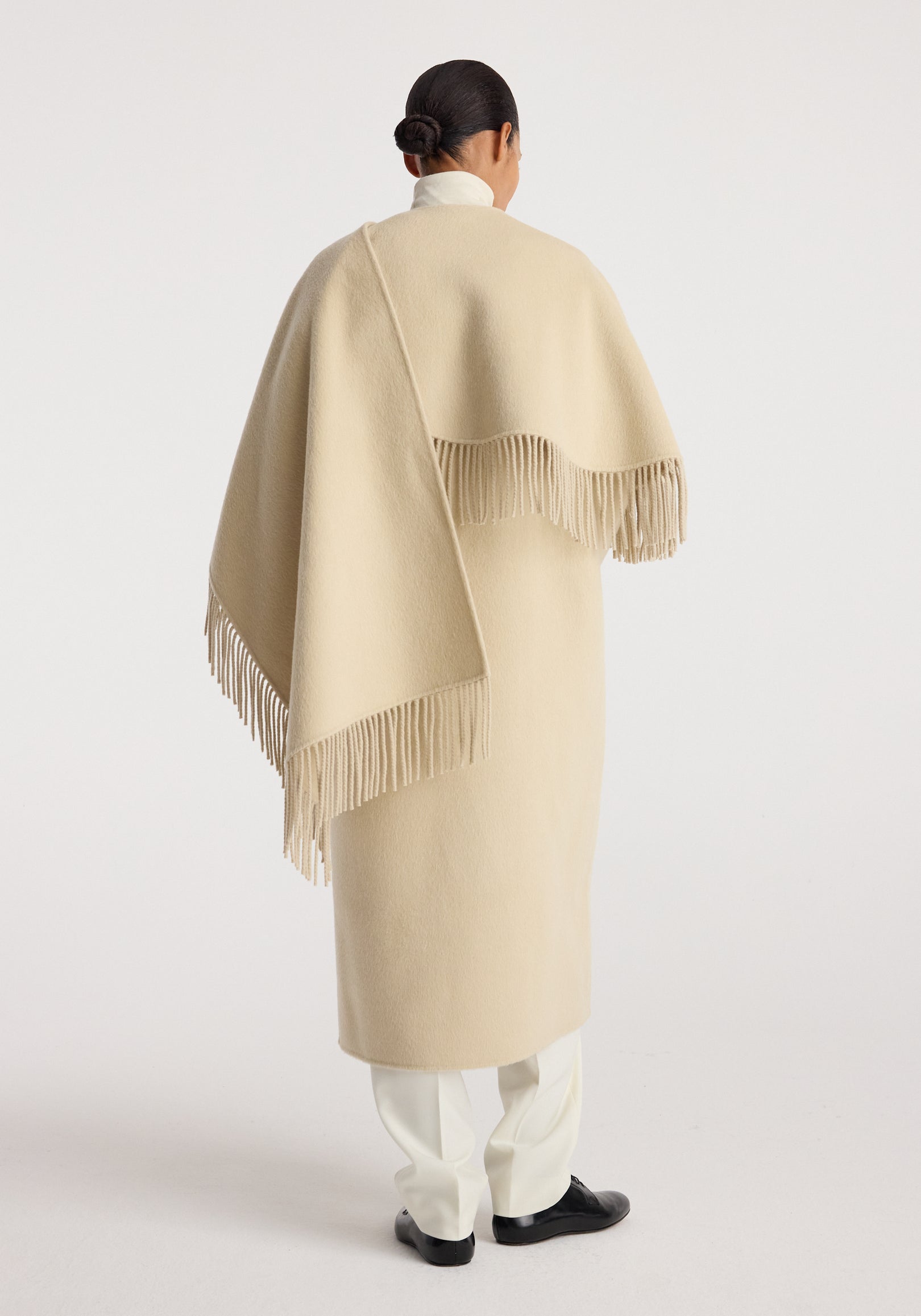 Back view of a person wearing a cream-colored Double-faced fringe coat | butter with long fringes along the edges, paired with white pants and black shoes against a plain background.