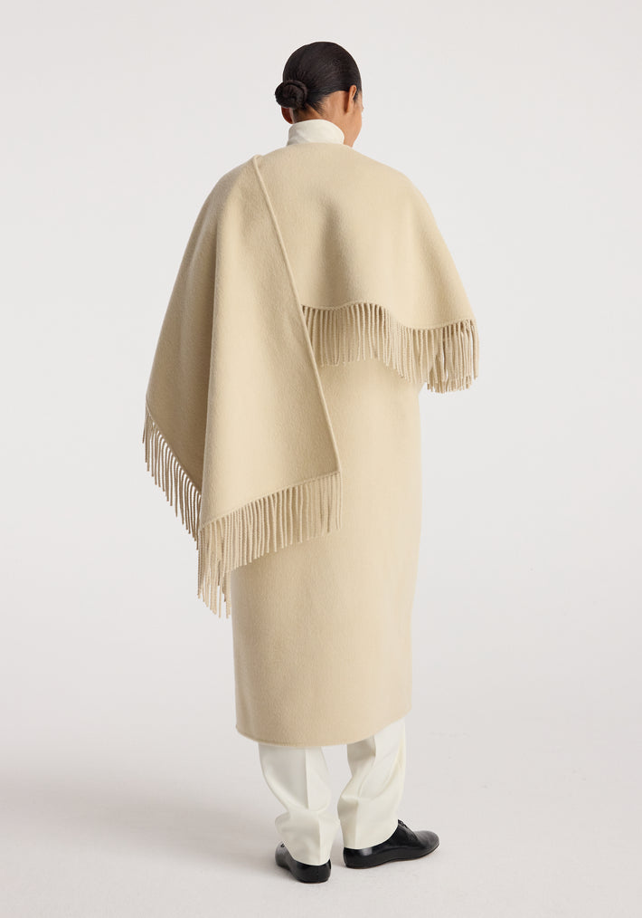 Back view of a person wearing a cream-colored Double-faced fringe coat | butter with long fringes along the edges, paired with white pants and black shoes against a plain background.