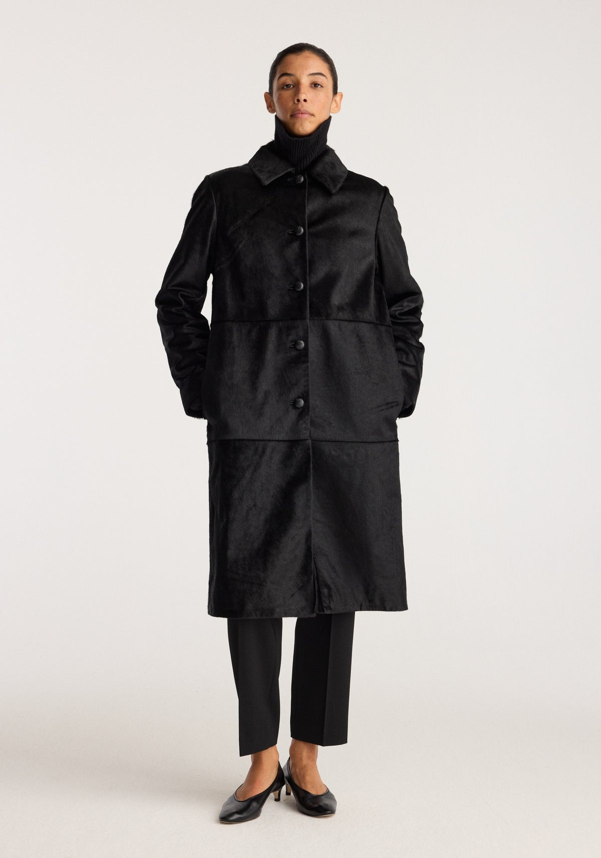 Black ponyhair coat | black worn by a model with a button front collar long sleeves side pockets and knee length silhouette styled with black trousers and flats