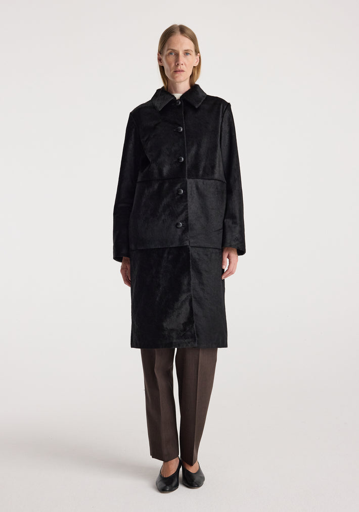 Woman wearing a long black coat with a matte finish, buttoned down the front, paired with dark trousers and black shoes, Black ponyhair coat | black.