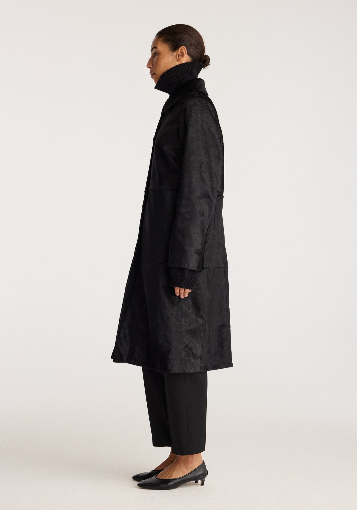Black ponyhair coat | black shown in side profile on a model, featuring a high collar, long silhouette, and black trousers with pointed shoes