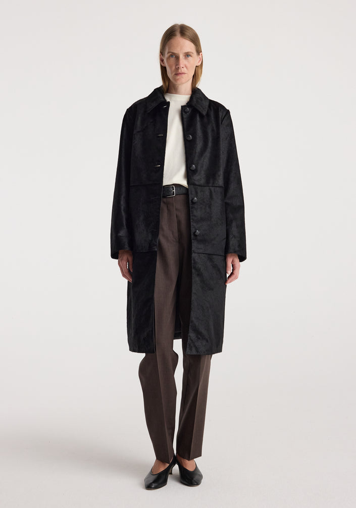 Woman wearing a long black coat with a matte finish, buttoned down the front, paired with dark trousers and black shoes, Black ponyhair coat | black.