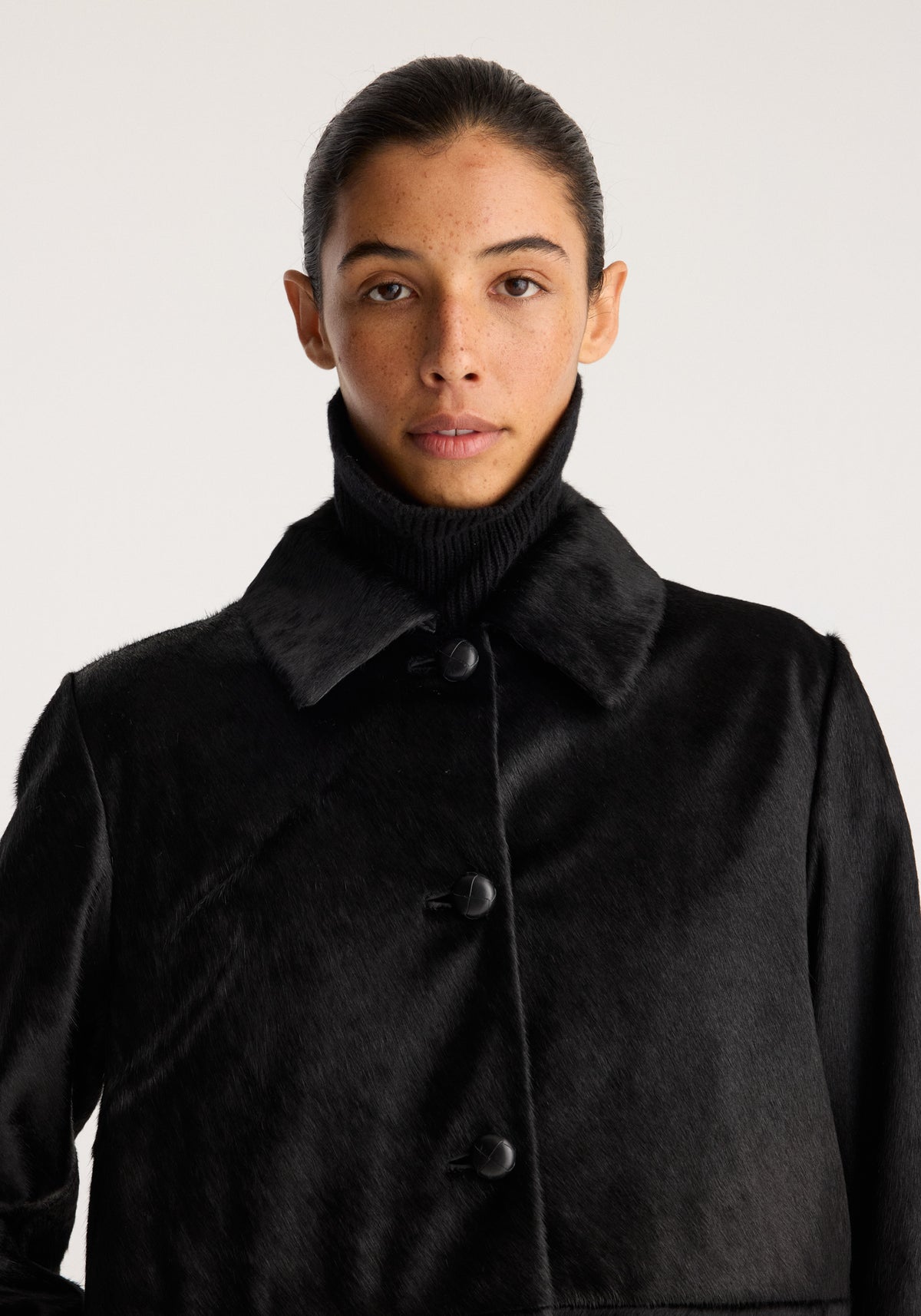 Black ponyhair coat | black shown on a model with a plush textured finish high collar front buttons and long sleeves against a neutral background