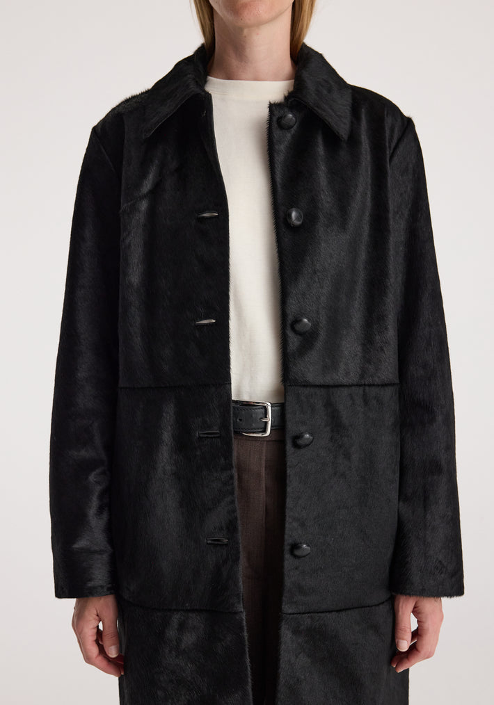 Close-up of a person wearing a sleek Black ponyhair coat | black with button closures, collar, and long sleeves over a white top and brown pants.