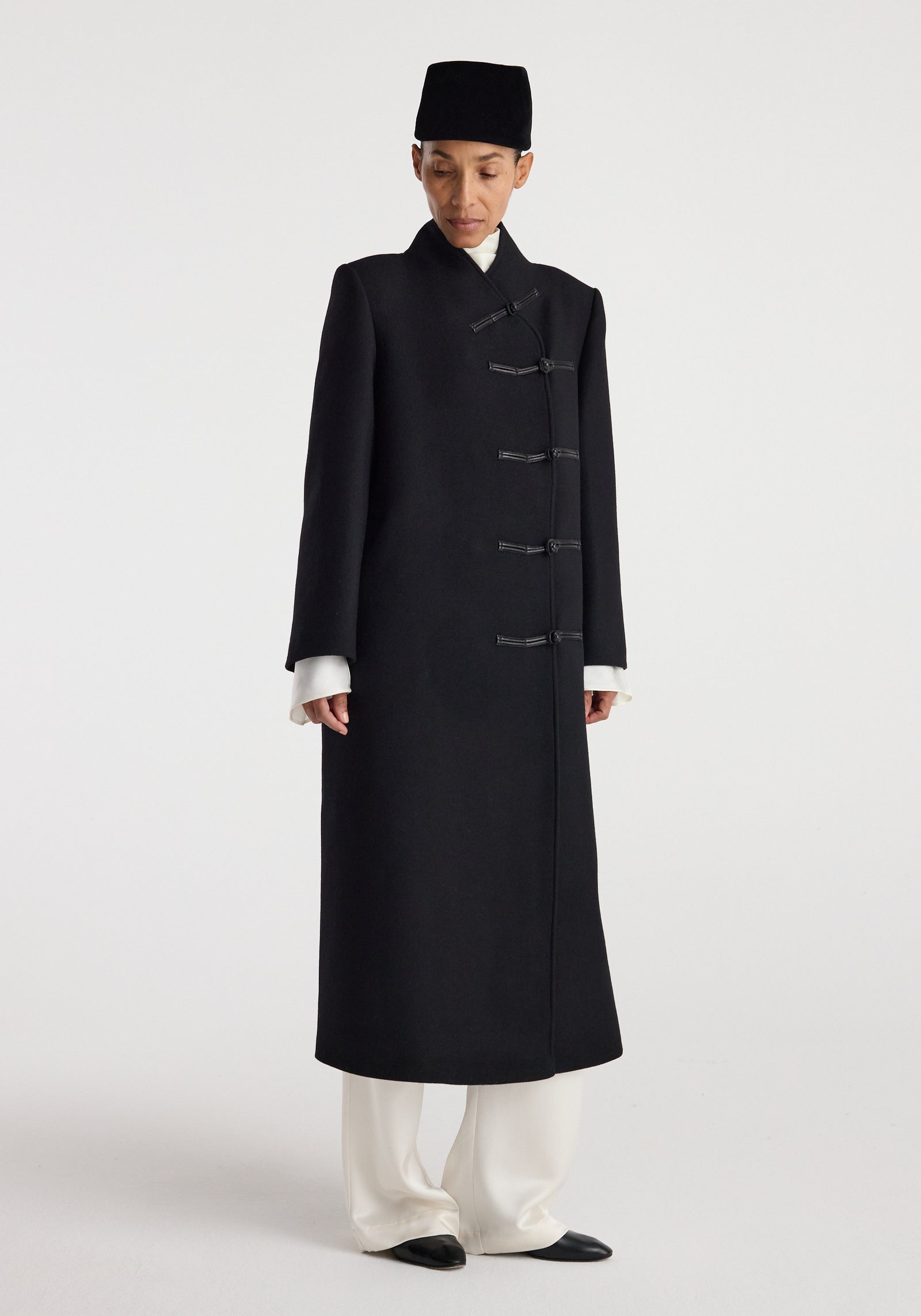 Woman wearing a long black Pankou closure wool coat | black with a stand collar, paired with white wide-leg pants, black shoes, and a black hat, standing against a plain background.