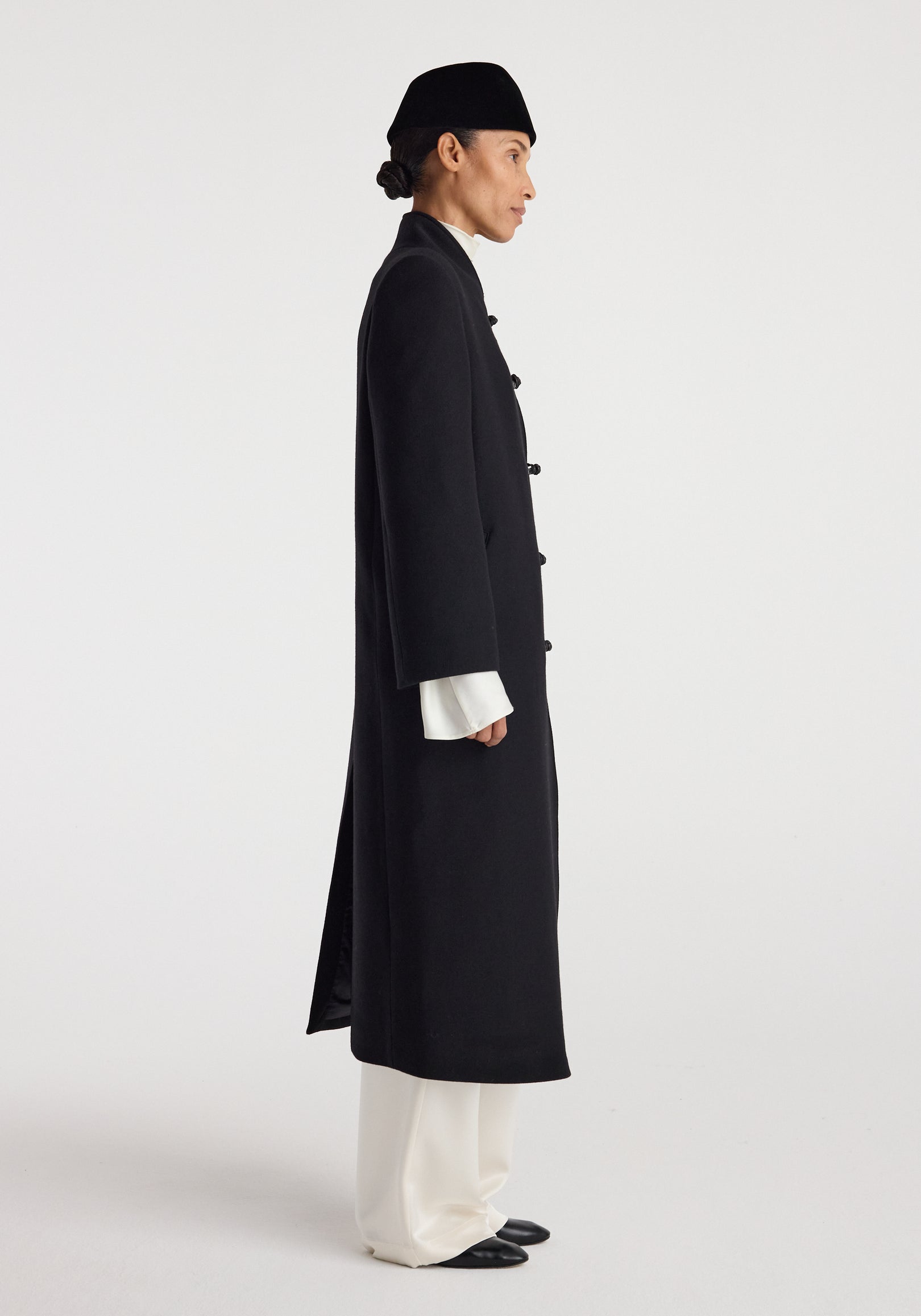 Side view of a person wearing a long black Pankou closure wool coat | black with traditional knot buttons, paired with loose white trousers and a black hat against a plain background.
