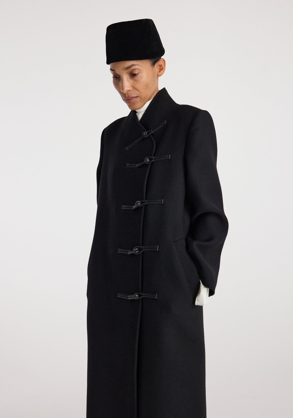 Model wearing a black Pankou closure wool coat | black with traditional toggle fastenings, standing against a plain light background with hands in pockets and a contemplative expression.
