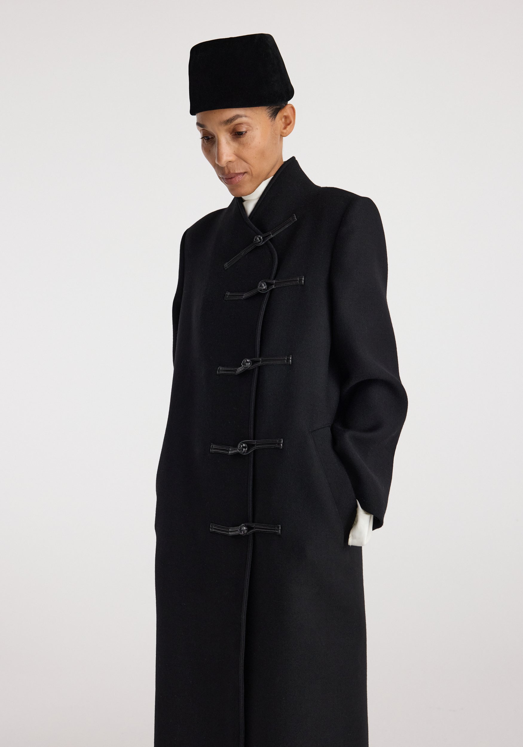 Model wearing a black Pankou closure wool coat | black with traditional toggle fastenings, standing against a plain light background with hands in pockets and a contemplative expression.