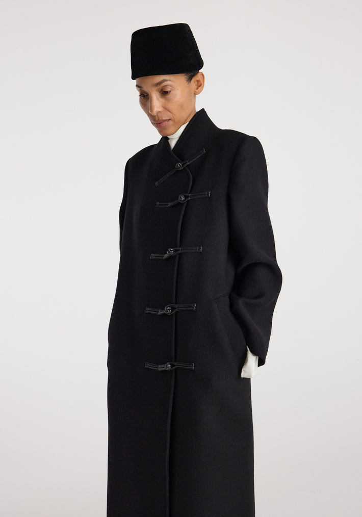 Model wearing a black Pankou closure wool coat | black with traditional toggle fastenings, standing against a plain light background with hands in pockets and a contemplative expression.