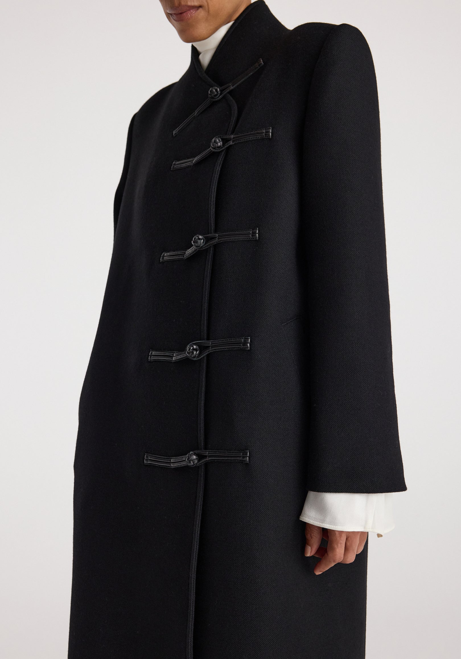 Close-up of a person wearing a black Pankou closure wool coat | black featuring traditional knotted fastenings and a high collar over a white turtleneck.