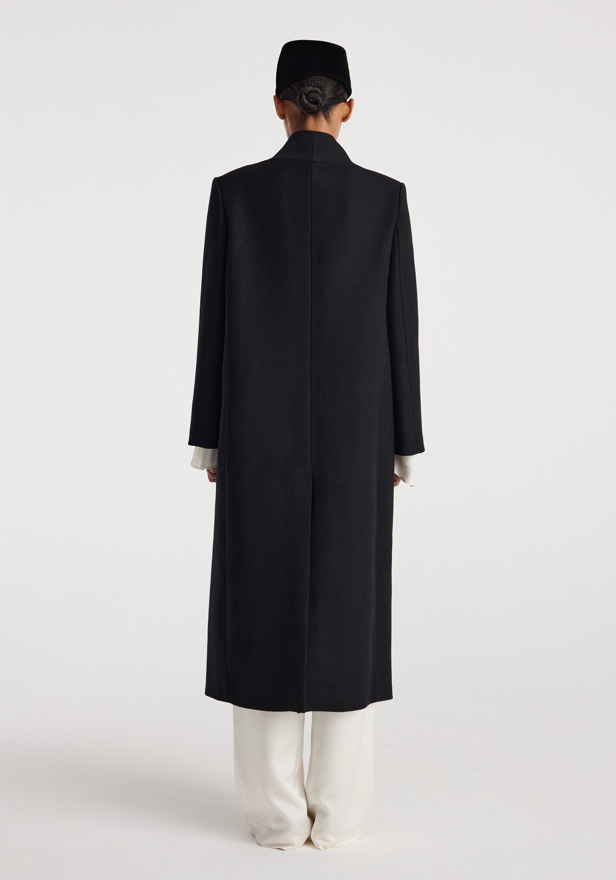 Back view of a person wearing a long black Pankou closure wool coat | black over white pants against a plain white background.