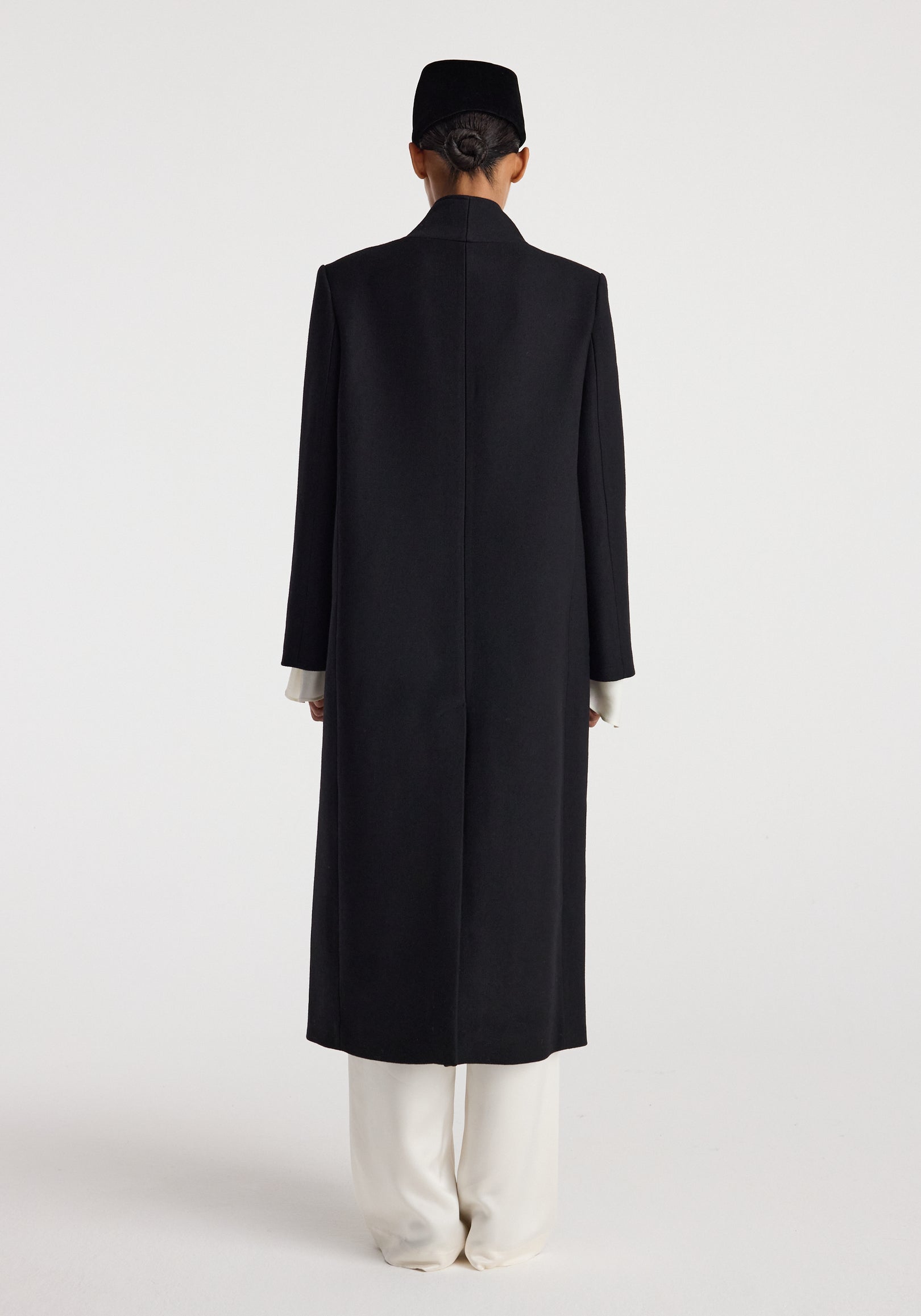 Back view of a person wearing a long black Pankou closure wool coat | black over white pants against a plain white background.