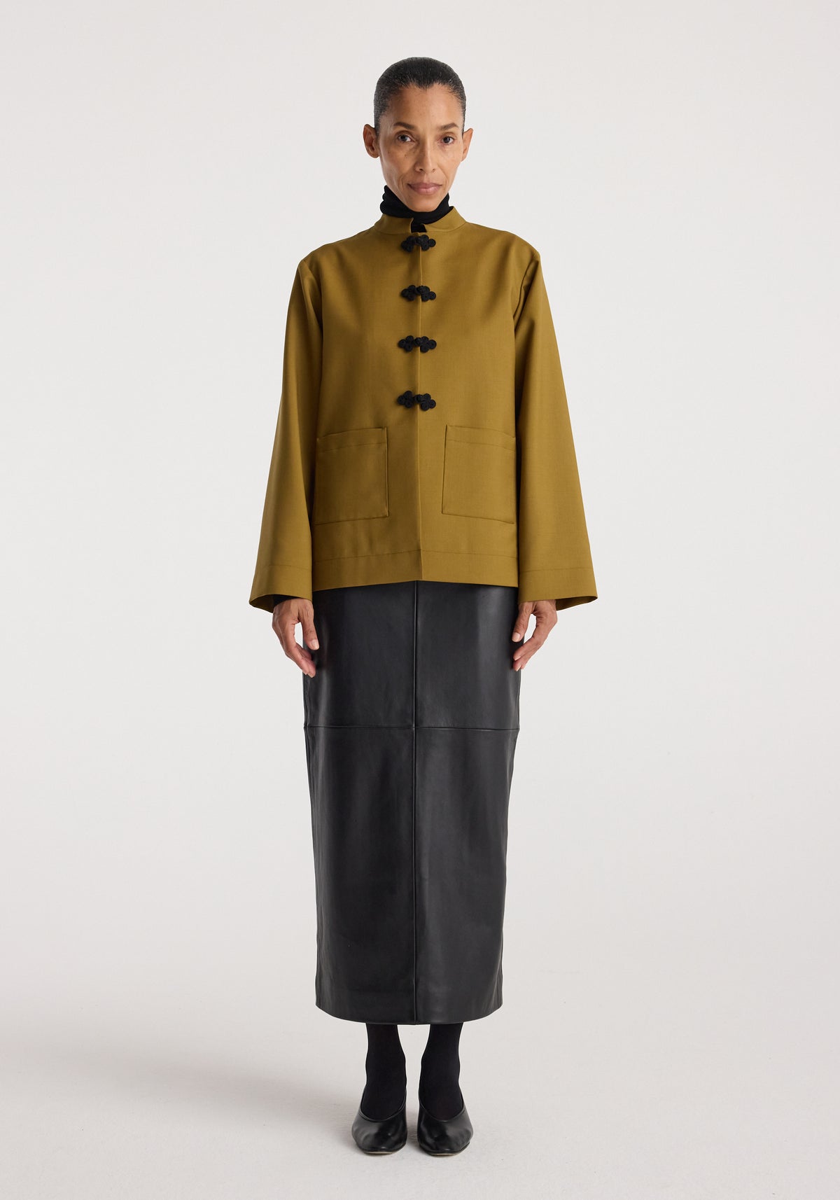 Woman wearing a Pankou closure jacket | mustard with black traditional knot buttons, paired with a long black skirt and black shoes, standing against a plain background.