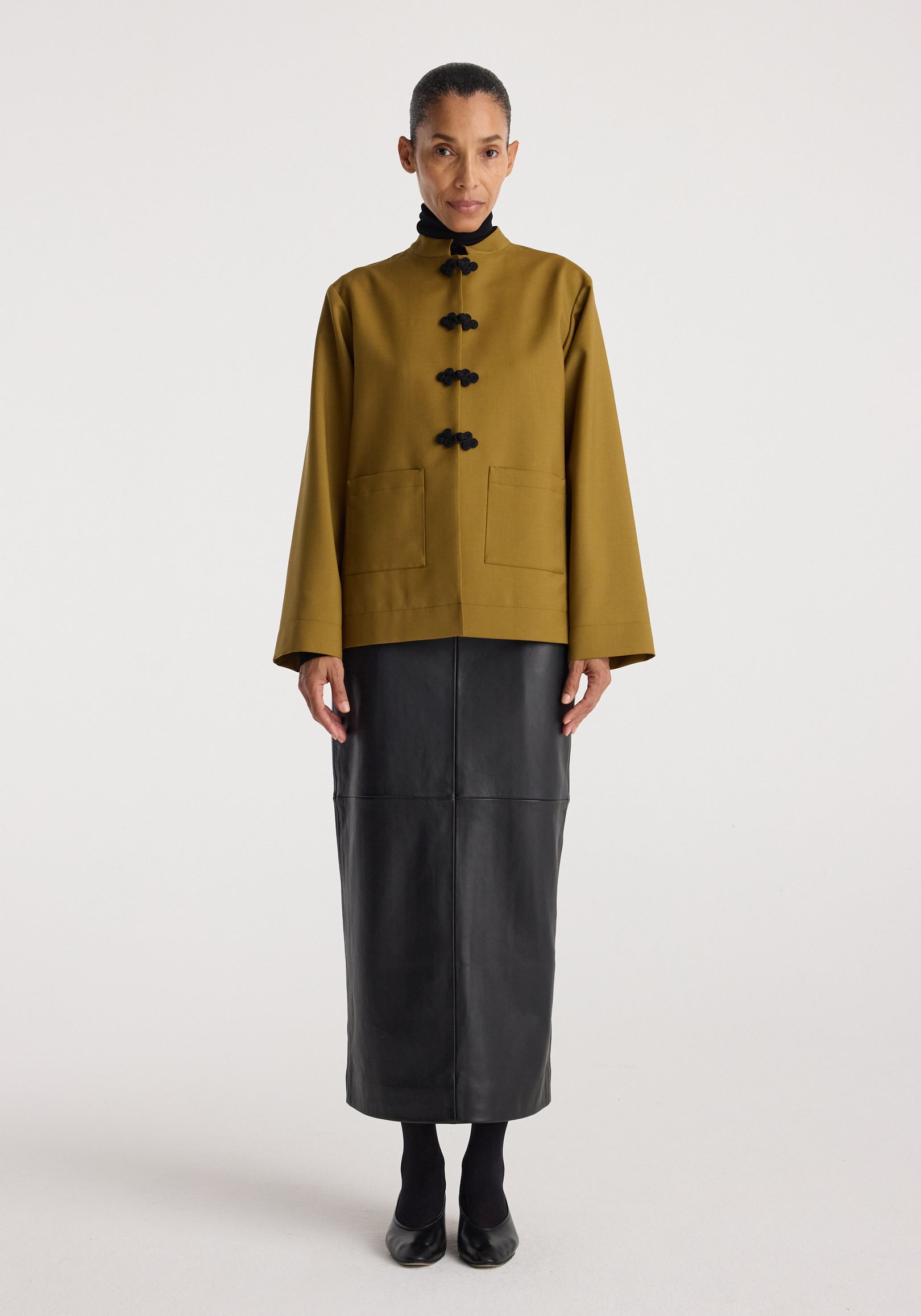 Woman wearing a Pankou closure jacket | mustard with black traditional knot buttons, paired with a long black skirt and black shoes, standing against a plain background.