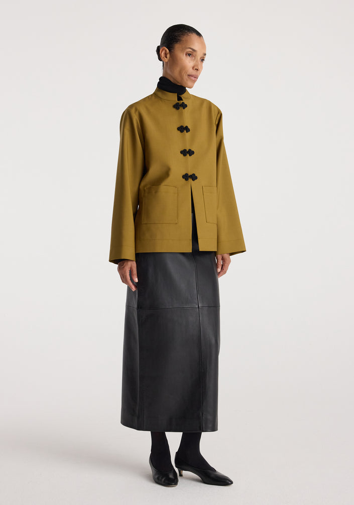 Woman wearing a Pankou closure jacket | mustard with black traditional knot buttons, paired with a long black skirt and black shoes, standing against a plain background.