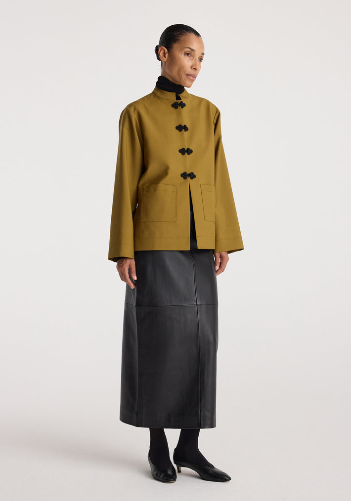 Woman wearing a mustard Pankou closure jacket with black toggle buttons, paired with a black turtleneck and a long black leather skirt against a plain background.