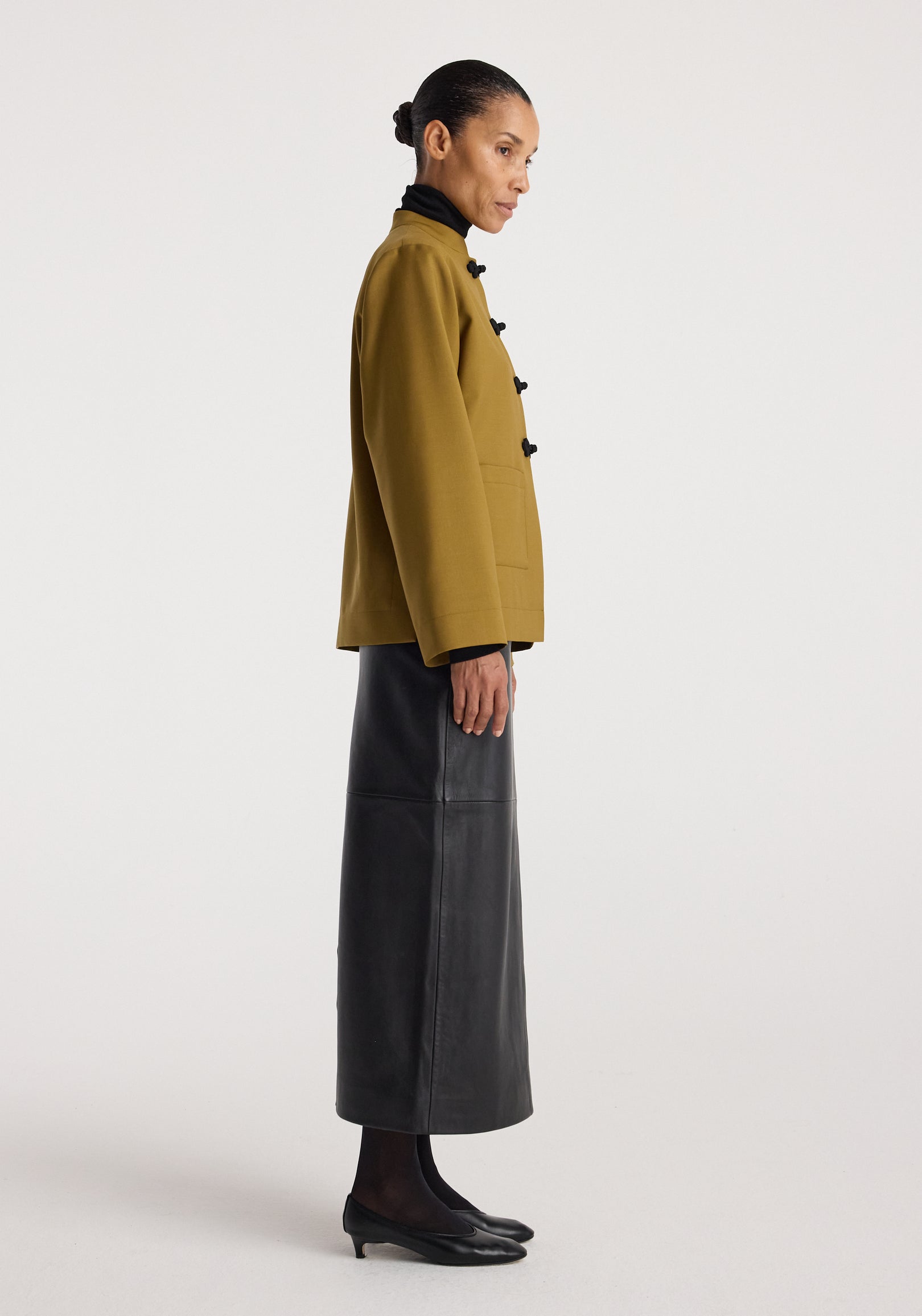 Woman wearing a mustard Pankou closure jacket with side toggle buttons, paired with a long black leather skirt and black low-heeled shoes against a plain background.
