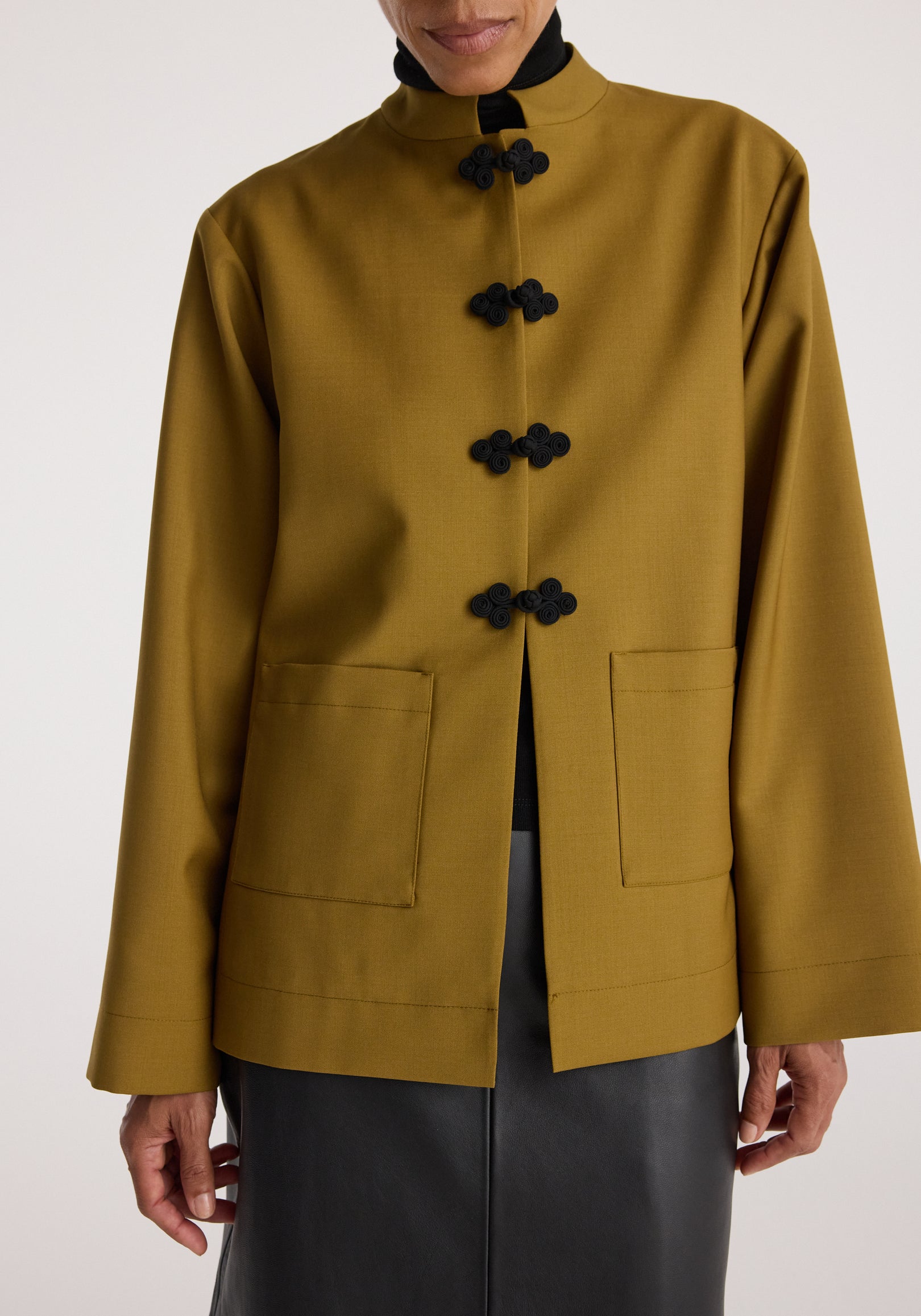 Woman wearing a mustard-colored jacket with Pankou closure featuring black frog buttons, patch pockets, and a high collar over a black top and skirt, Pankou closure jacket | mustard.
