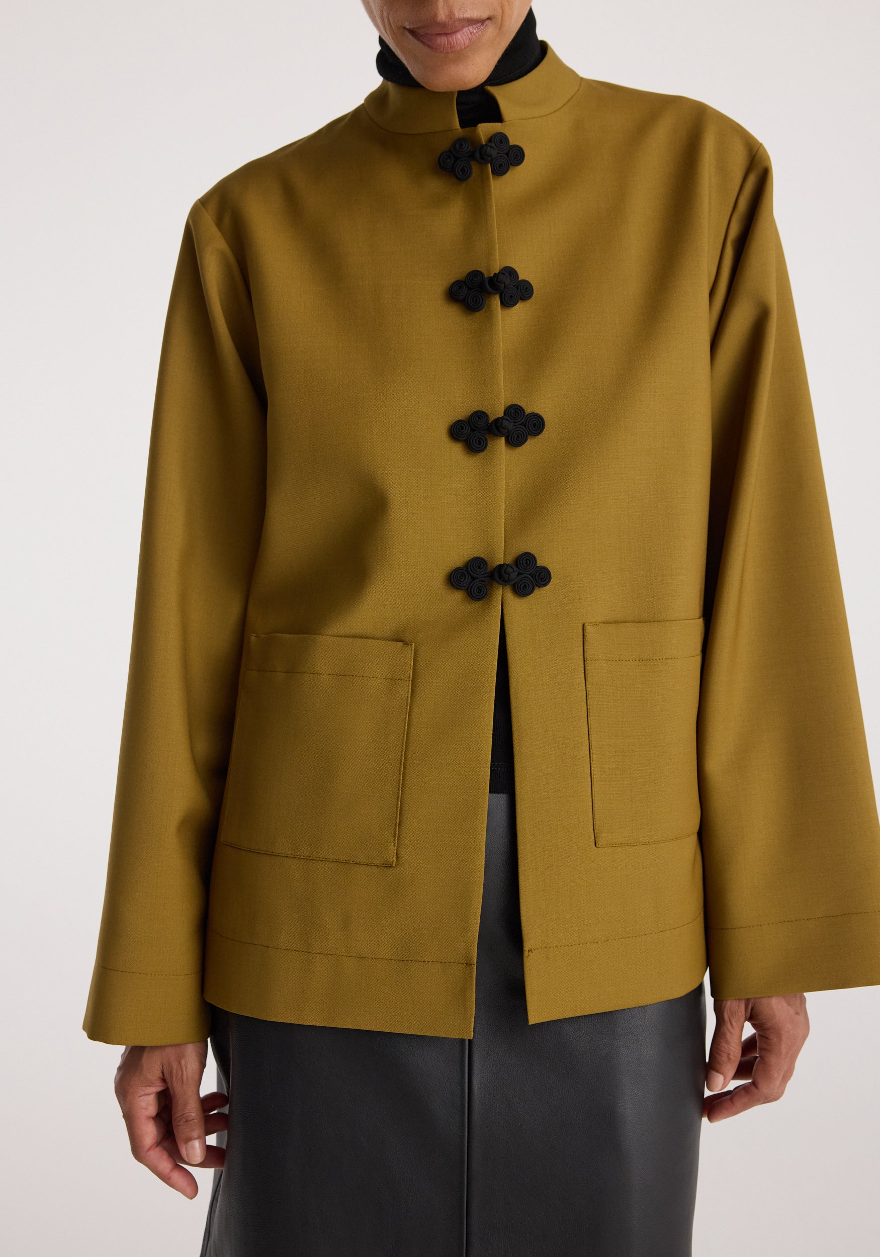 Woman wearing a mustard-colored jacket with Pankou closure featuring black frog buttons, patch pockets, and a high collar over a black top and skirt, Pankou closure jacket | mustard.