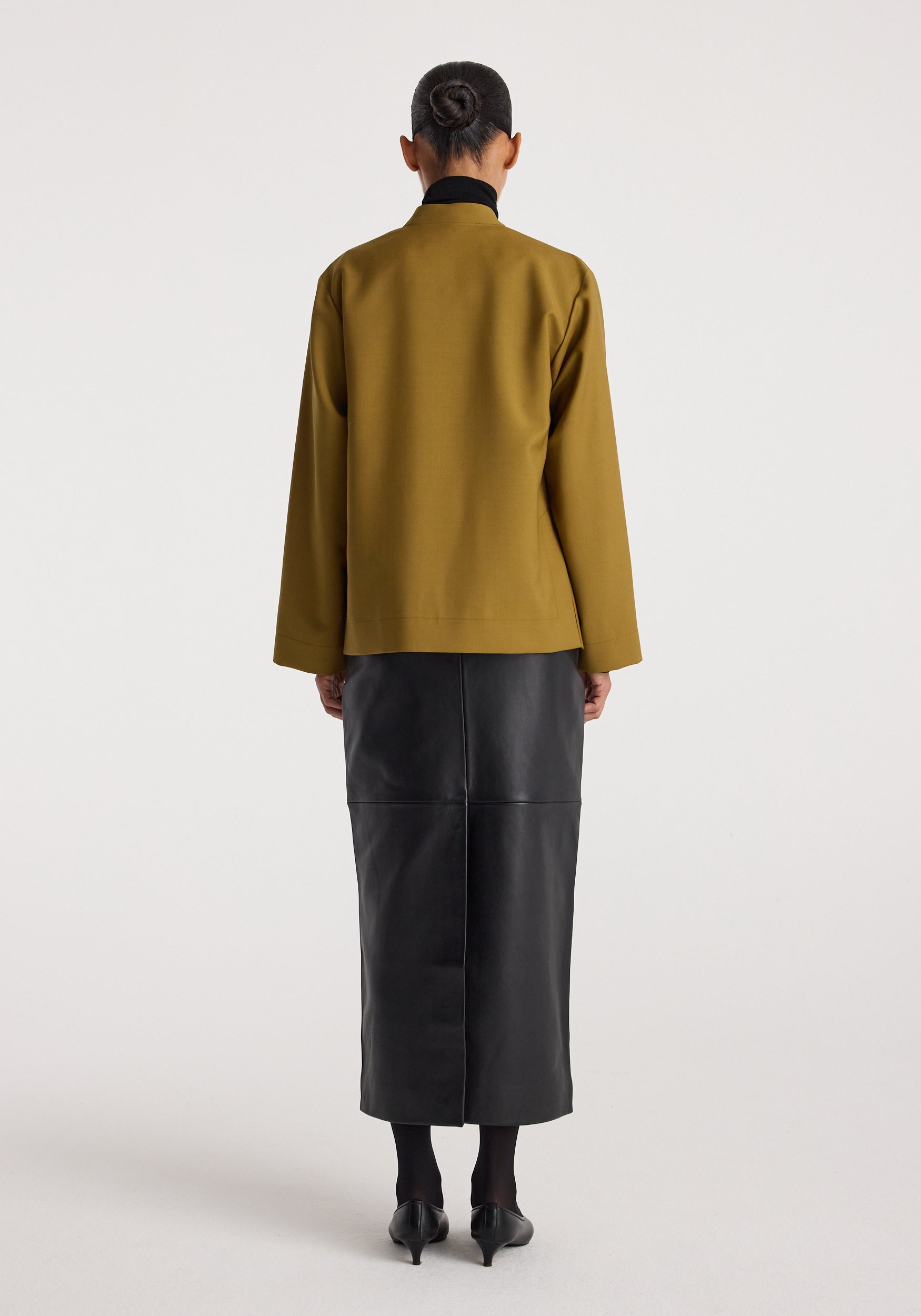 Woman wearing a Pankou closure jacket | mustard with long sleeves, paired with a black leather midi skirt and black heels, shown from the back against a plain white background.