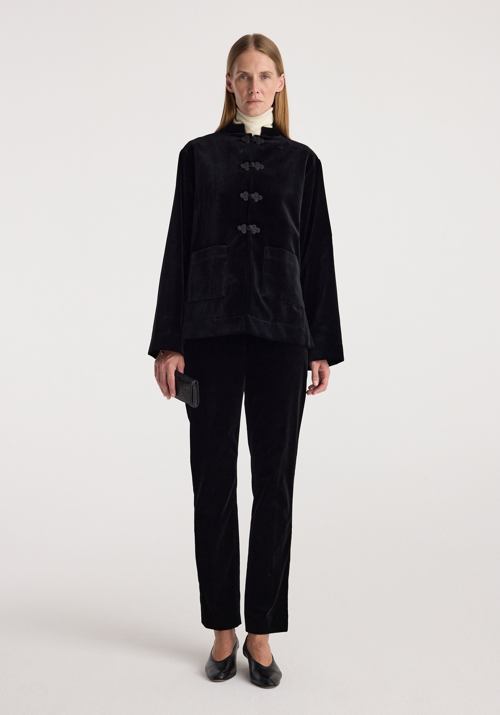 Woman wearing a black Pankou closure velvet jacket | black with a high collar, paired with matching black velvet pants, holding a small black clutch and wearing black shoes.