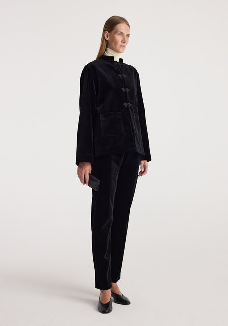 Woman wearing a black Pankou closure velvet jacket | black with a high collar, paired with matching black velvet pants, holding a small black clutch and wearing black shoes.