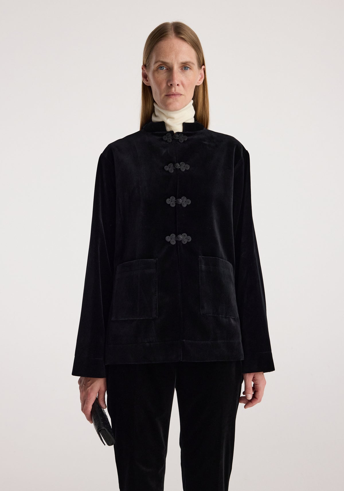 Woman wearing a black velvet jacket with pankou closure and large front pockets, paired with a white turtleneck and holding a small black clutch, minimalistic background.