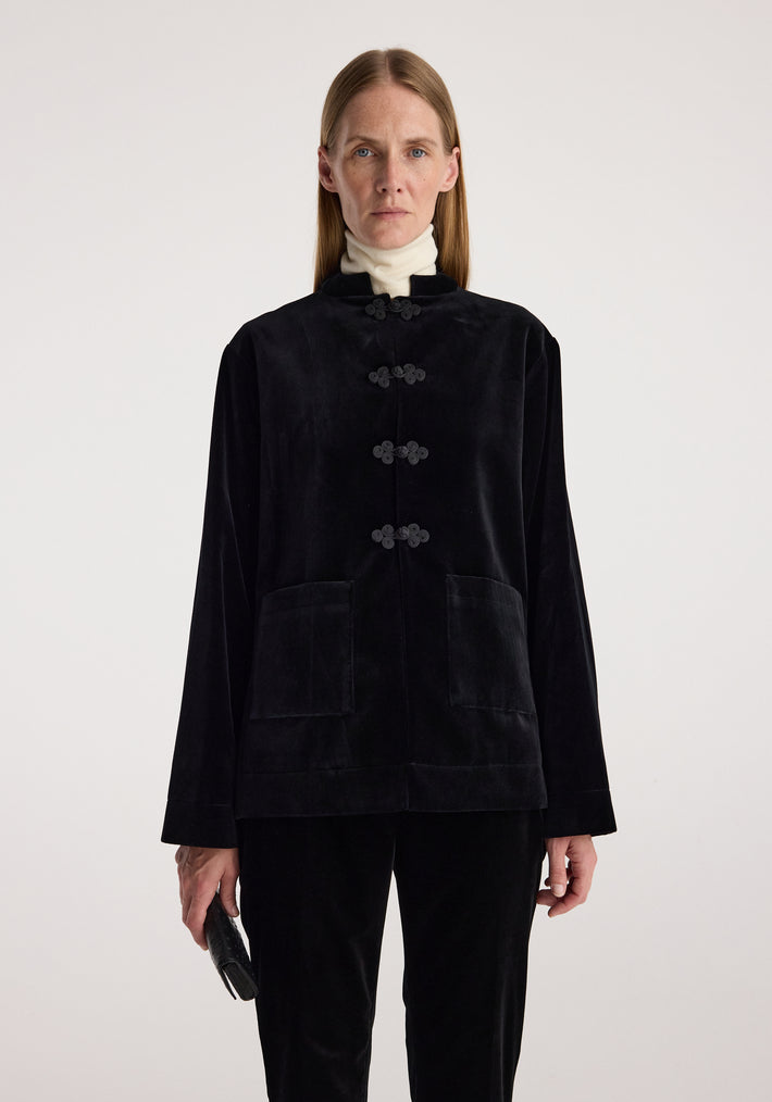 Woman wearing a black velvet jacket with pankou closure and large front pockets, paired with a white turtleneck and holding a small black clutch, minimalistic background.