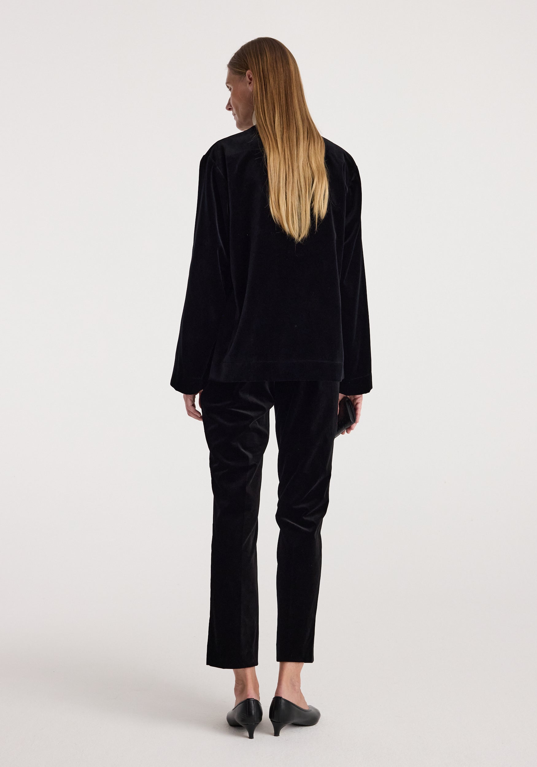 Woman wearing a black Pankou closure velvet jacket | black, shown from the back, paired with matching black velvet pants and black heeled shoes.