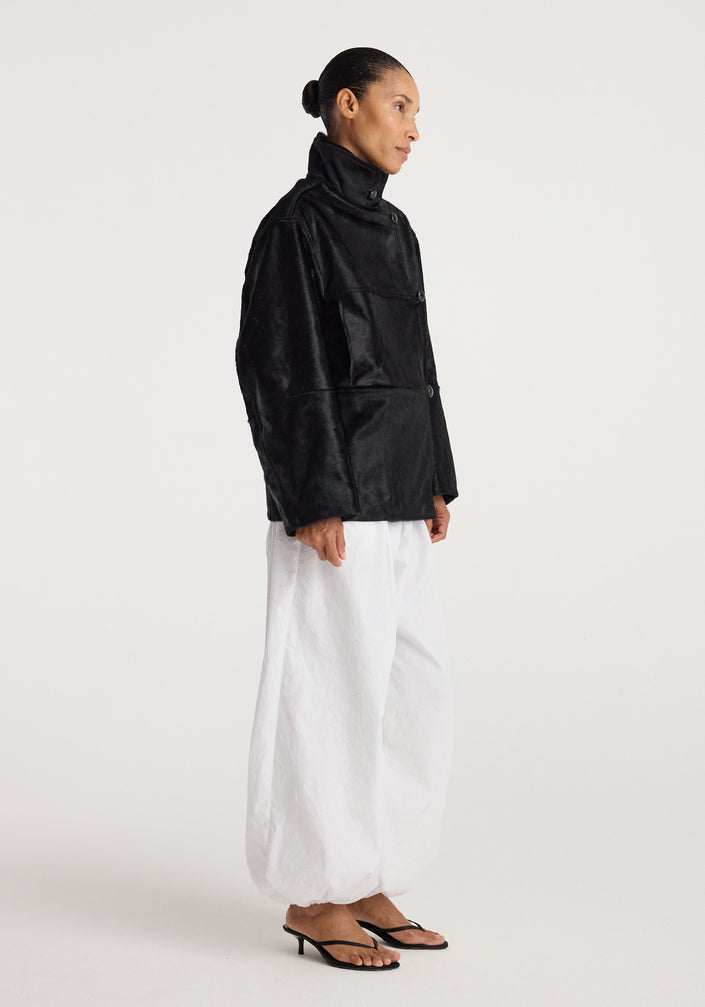 Woman wearing a black ponyhair jacket | black with a high collar and button closure, paired with loose white pants and black sandals against a plain background.
