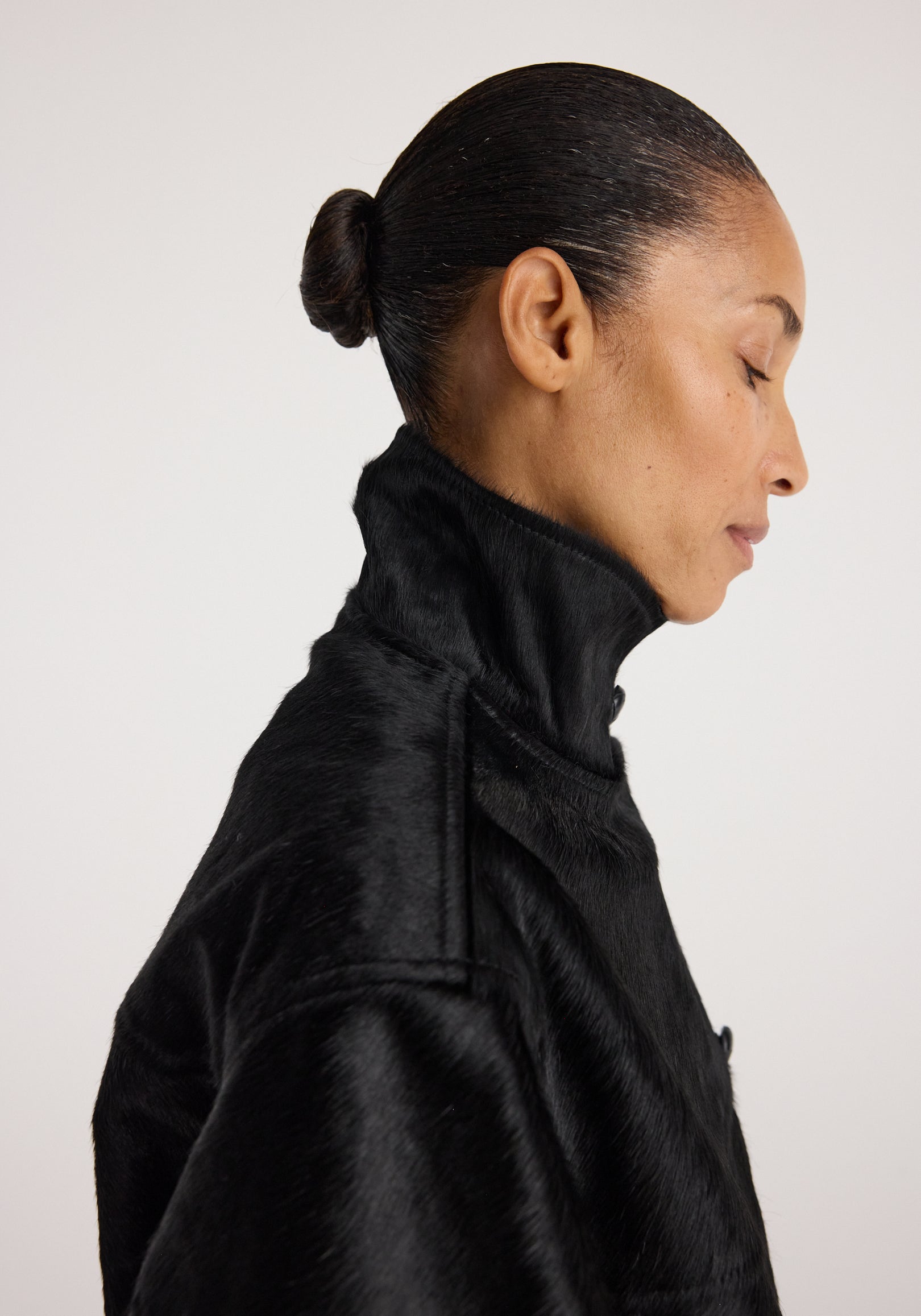 A woman with a sleek bun hairstyle wearing a high-collar black ponyskin jacket, showcasing the texture and luxurious finish of the Black ponyhair jacket | black.